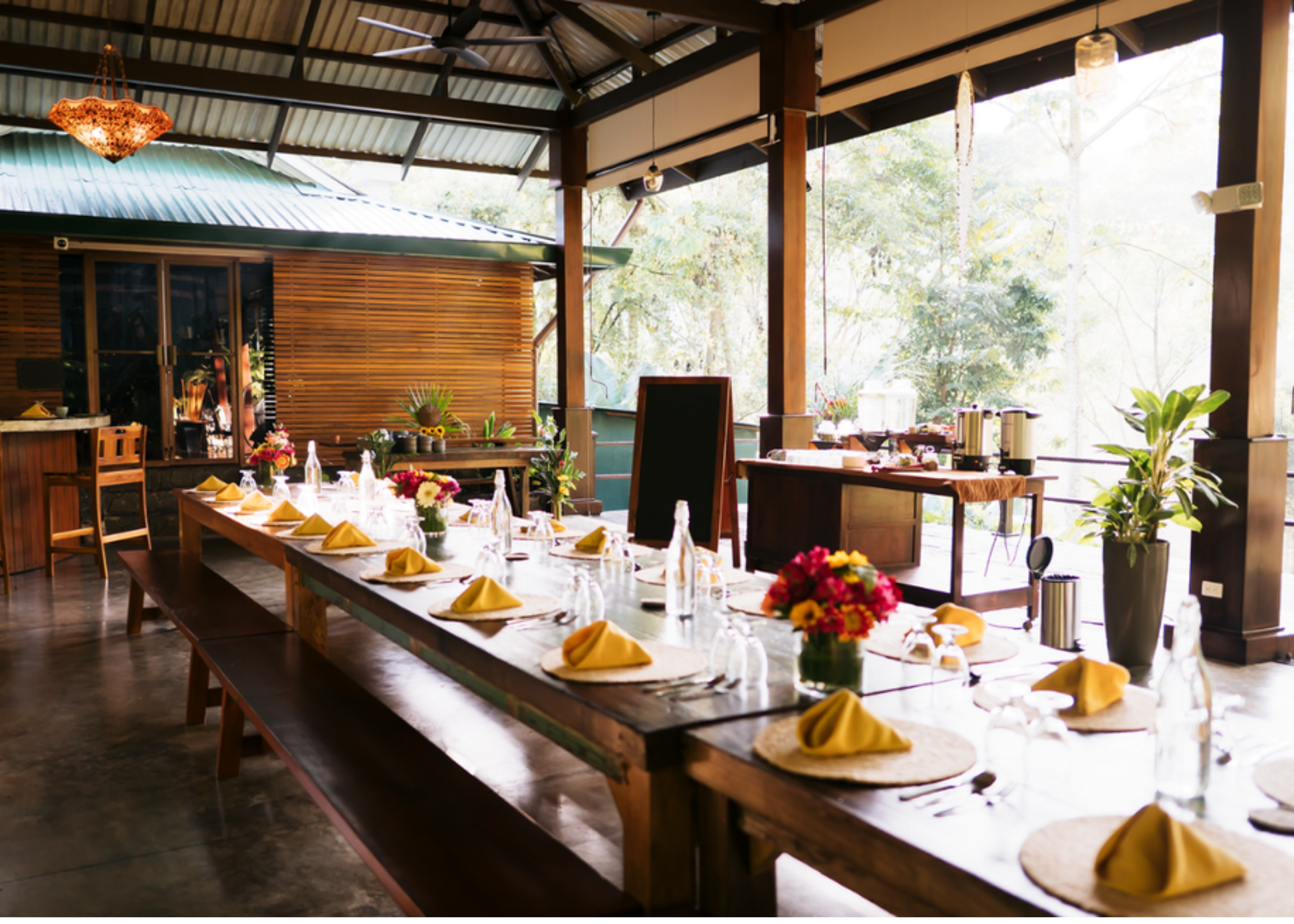 Open air dining area with a long table set for dinner with round placemats, yellow napkins, silverware, and water bottles. Lots of plants around.