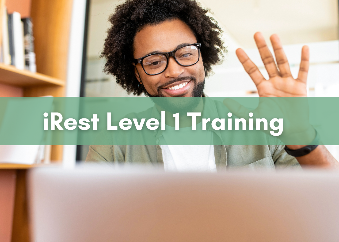 A smiling man wearing glasses waves at his laptop screen while participating in an online meeting or training. A green banner across the image reads “iRest Level 1 Training” in bold white text.